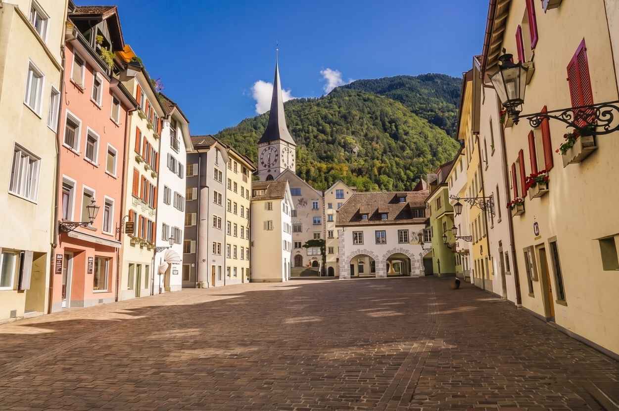 Blick durch einen historischen Holzbogen auf eine schmale Kopfsteinpflastergasse in der Churer Altstadt, mit Fußgängern, traditionellen Gebäuden, Blumenkübeln, einer antiken Wandlaterne und einem hängenden Holzschild mit der Aufschrift „Zum Stern Cobblestone Alley“.